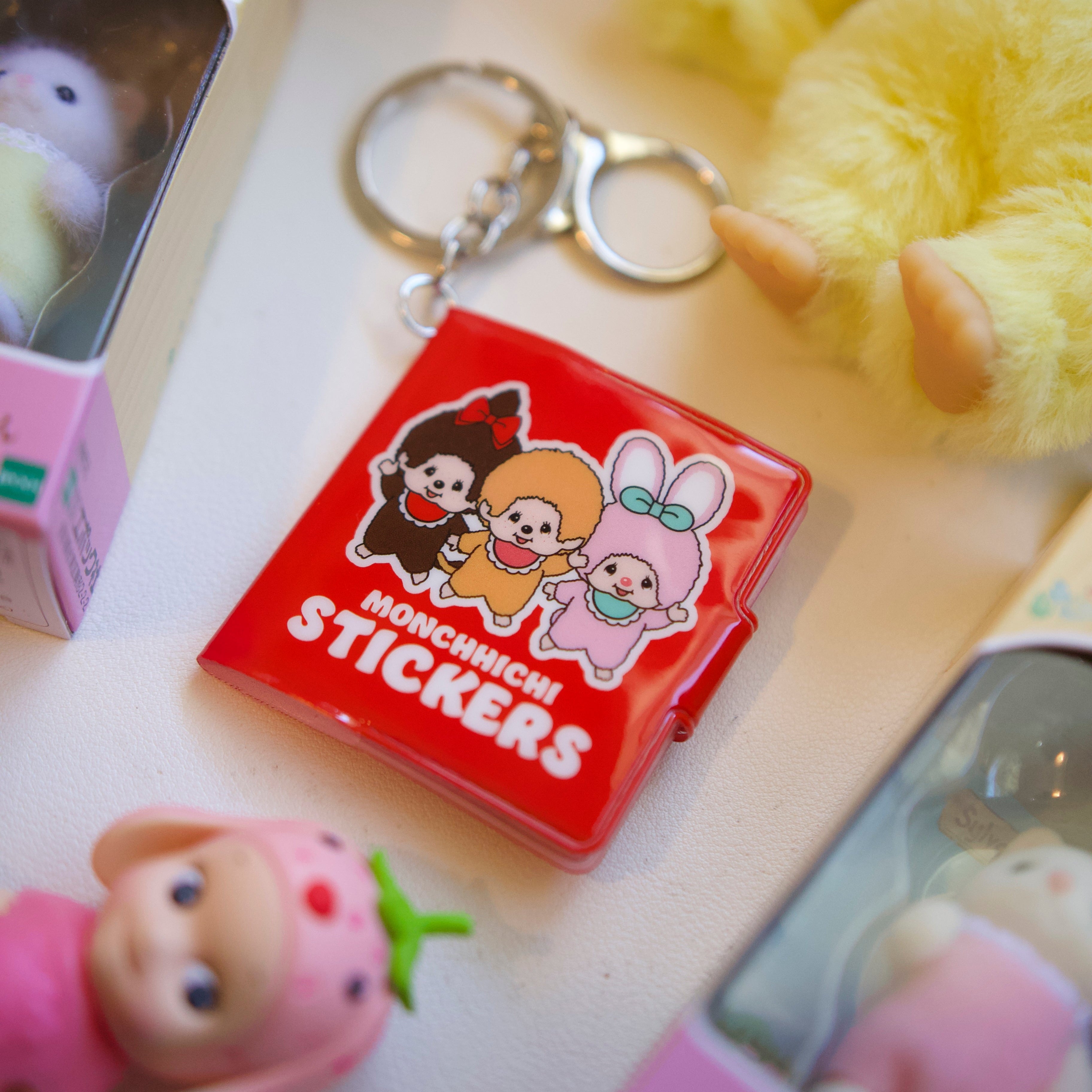Monchhichi mini sticker book keychain by Pipsticks — red cover with kawaii character art, shown as a bag charm with cute collectible figurines