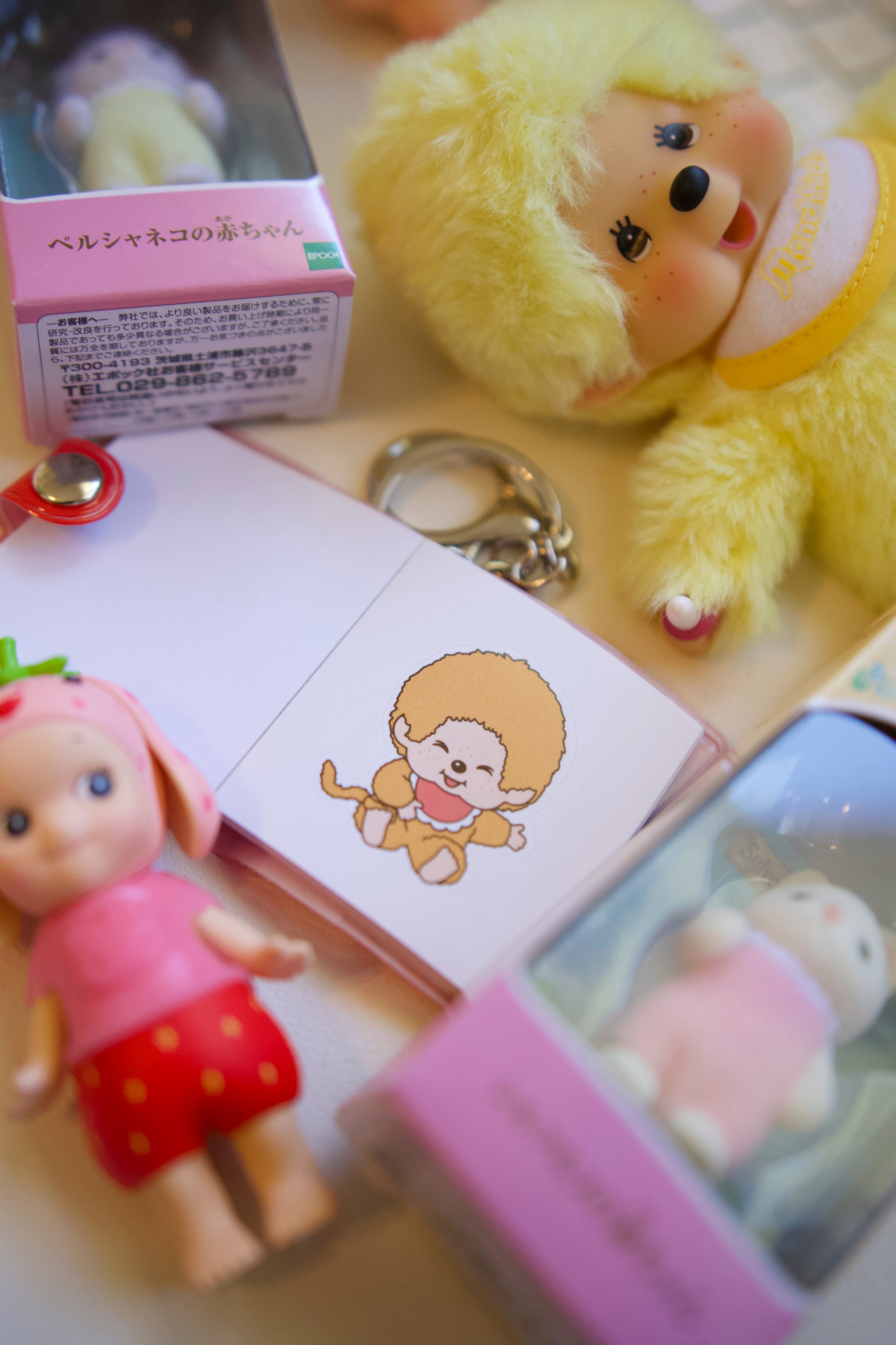 Open Monchhichi mini sticker book keychain showing kawaii character sticker pages, surrounded by cute collectible figurines and toys